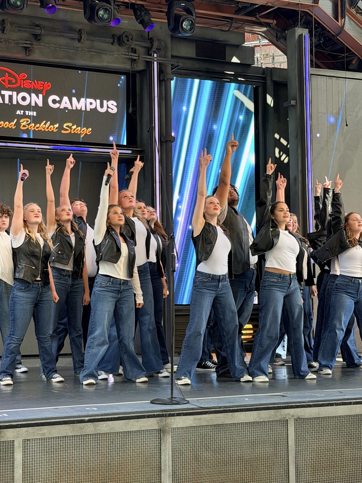 Aspire Young Artists mid-number on the Hollywood Backlot Stage, arms raised in unison