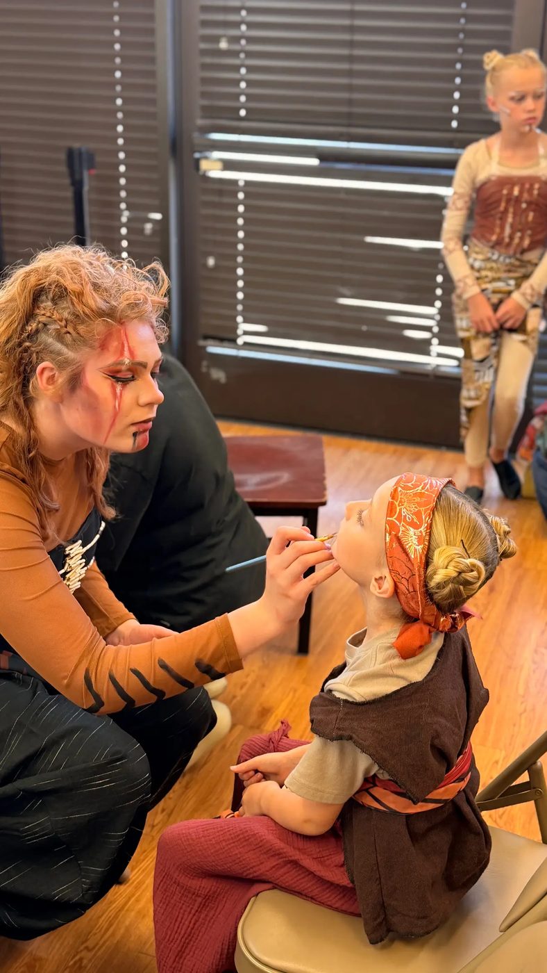 A costumed Young Artist paints face makeup on a younger castmate