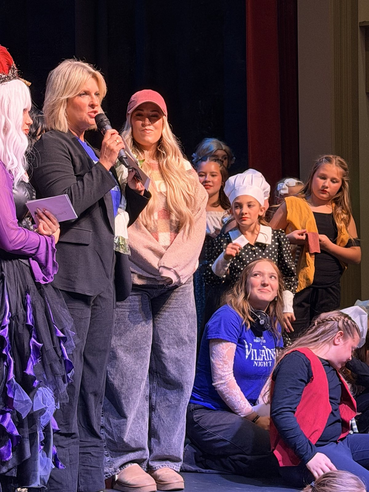 An Encore leader speaking into a microphone on stage surrounded by the Villains Night company