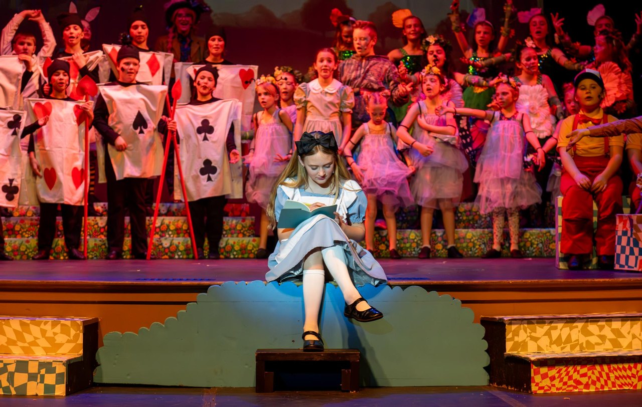 A young performer as Alice reads on stage while the full company performs behind her in Encore's Alice in Wonderland